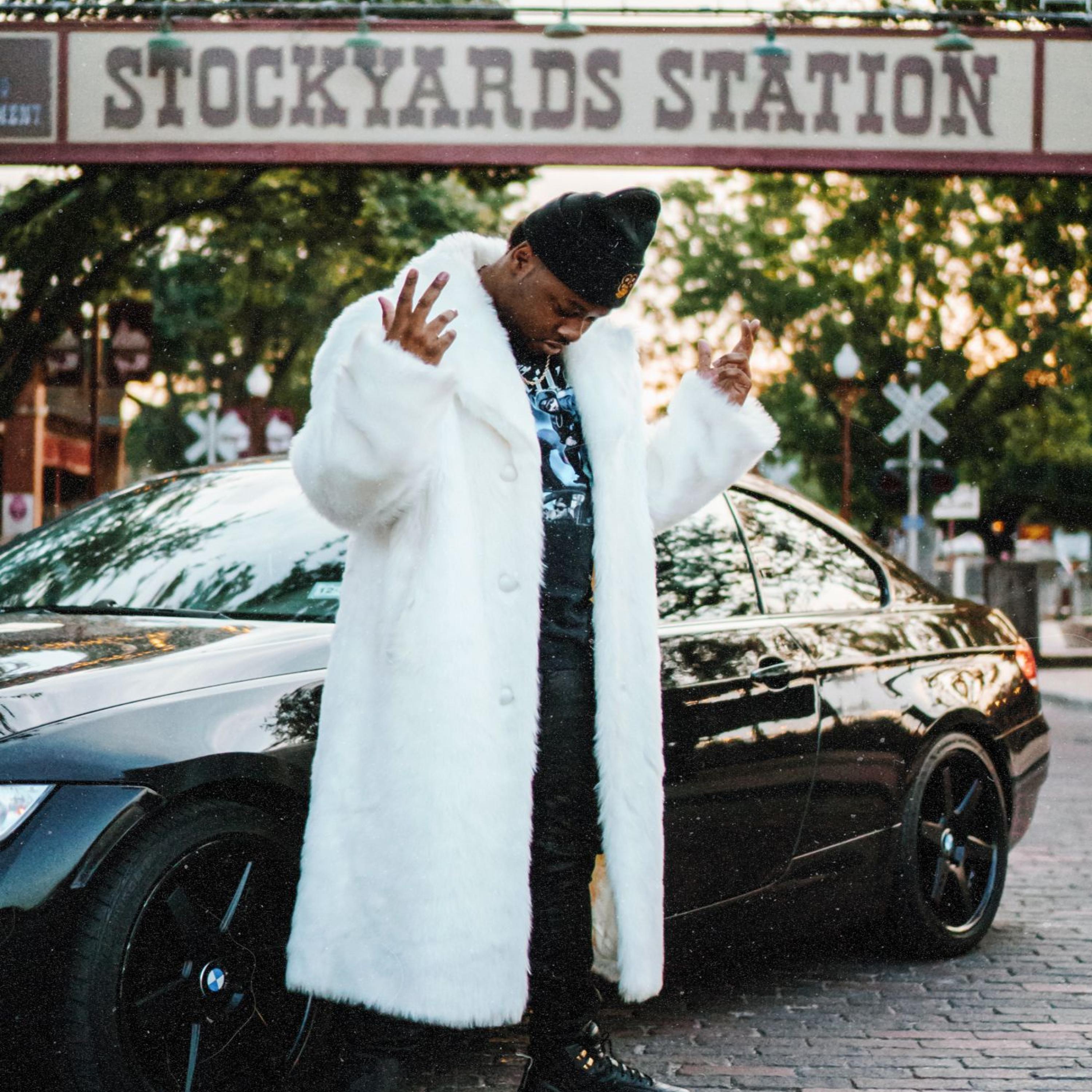 88 Killa wearing a white fur coat beside a black car at Stockyards Station in Fort Worth