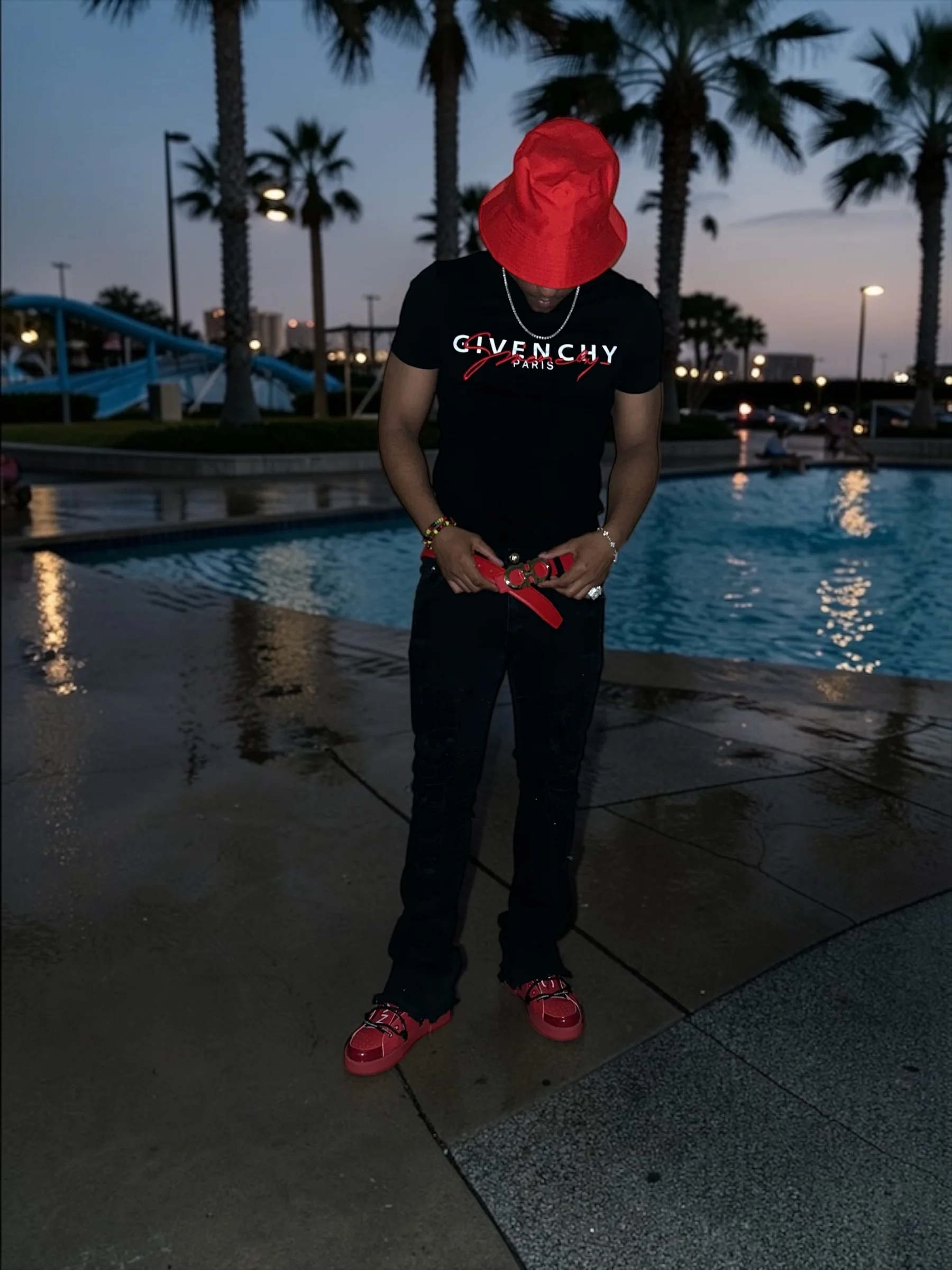 Doe Bwoy in a red bucket hat and black shirt beside a palm-lined swimming pool