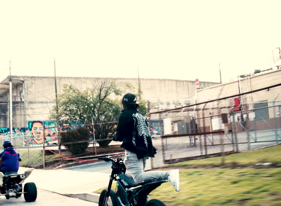 Fox BD on a dirt bike passing an outdoor fenced industrial area in the official video.